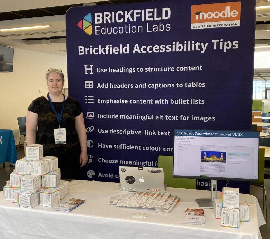 Karen Holland (Brickfield) standing in front of the Brickfield Accessibility Tips Banner on our stand. On the table in front is a some pile of Accessibility Tip Cubes, some leaflets, some business cards and a laptop with an external screen showing the demo of the Accessibility Toolkit Wizard.