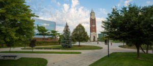 A scenic view of Oakland University campus featuring a tall clock tower with a brick facade and white base, surrounded by modern buildings, green lawns, and trees under a partly cloudy sky.