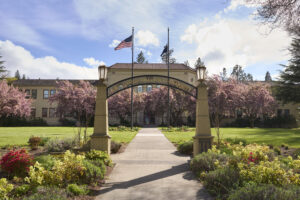 The image shows a picturesque entrance into a campus building, featuring a prominent archway with the words “Southern Oregon University” inscribed on it. Two stone pillars with lanterns stand on either side of the archway. The pathway leading to the building is bordered with gardens with a variety of flowers and plants. Behind the arch, there is a large, stately building with a tiled roof and multiple windows, framed by blooming cherry blossom trees. Two flag poles, with one representing the United States, are near the entrance. The sky above is partly cloudy, allowing sunlight to cast upon the scene.