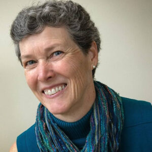 A woman with short, curly grey hair smiles warmly at the camera. She is wearing a teal top and a multicoloured scarf, with small earrings visible. The background is plain and light-coloured.