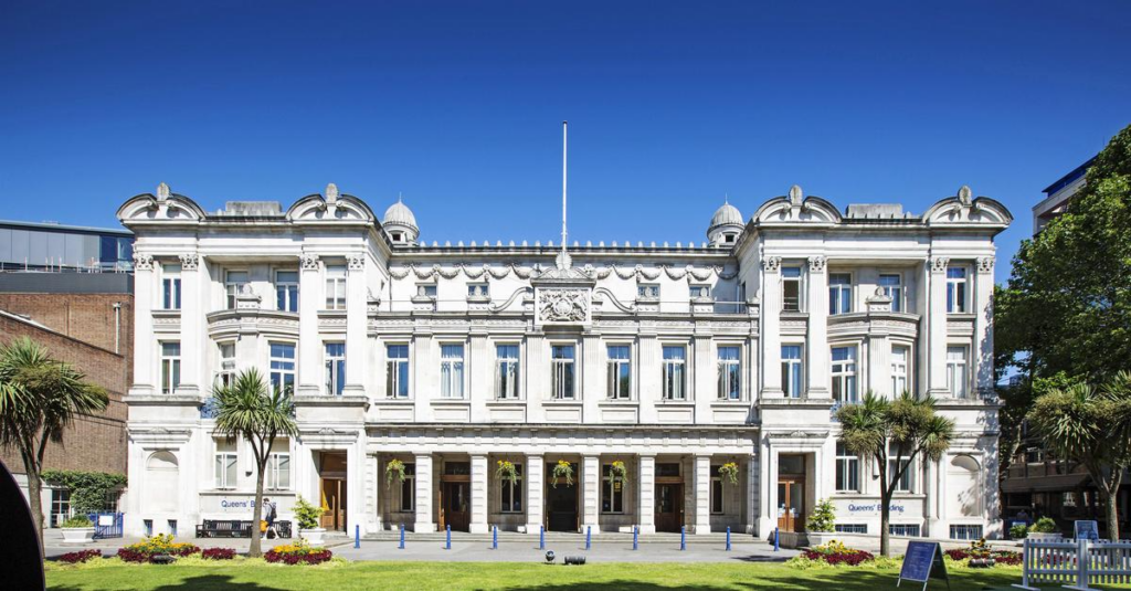 Front view of Queen Mary University, featuring a white building with tall columns, multiple windows and decorative elements on the roofline. The building is surrounded by greenery including palm trees and a lawn, under a clear blue sky.