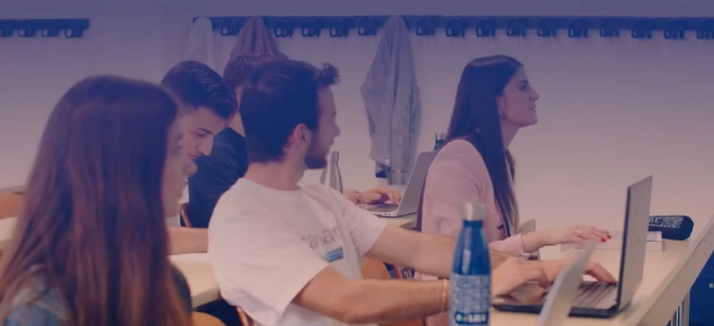 Students in a classroom at desks with their laptops looking towards a teacher who is not in the picture.