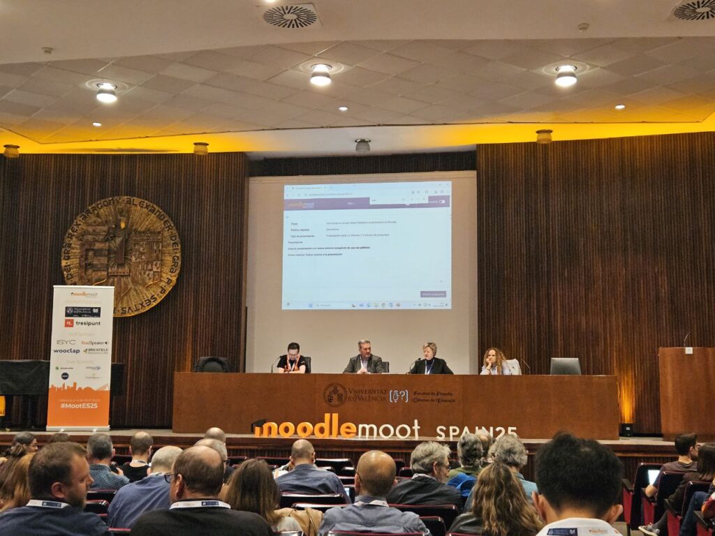 Moodlemoot Spain Welcome session in auditorium. A panel of three people sit at the top of the room in front of the screen with others watching from rows of chairs.