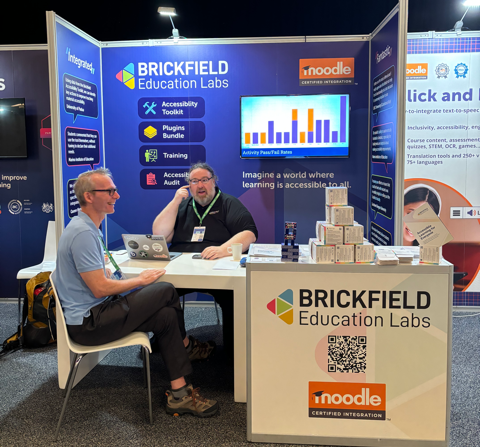 Exhibition booth for Brickfield Education Labs at MoodleMoot Global 2025, with Gavin Co-Founder of Brickfield Education Labs, and Fred Dixon sat at a table talking to each other. The table is in front of the booth setup, consisting of a TV and promotion of accessibility solutions.
