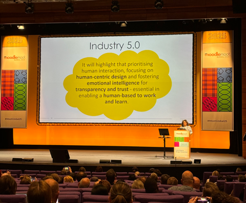 A large auditorium with attendees sat in seated rows facing a brightly lit stage. On the stage is a large screen which displays a slide titled "Industry 5.0". The slide reads: "It will highlight that prioritising human interaction, focusing on human-centric design and fostering emotional intelligence for transparency and trust - essential in enabling a human-based to work and learn". Dr Neema Pasha stands at the podium to the right of the stage.