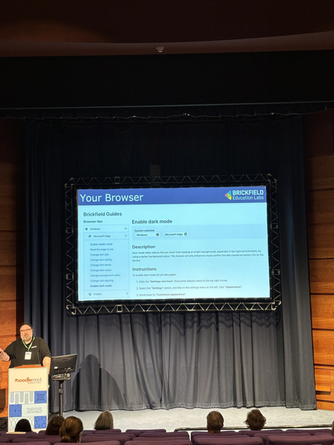 An auditorium with attendees in seated rows facing a brightly lit stage. On the stage is a large screen displaying a presentation slide titled "Brickfield Guides". The slide lists browser tips and accessible file options, such as configuring your browser, creating accessible documents and slides, and finding alternative formats. To the left of the screen, Gavin Henrick, Co-Founder of Brickfield Education Labs, stands at a podium presenting.