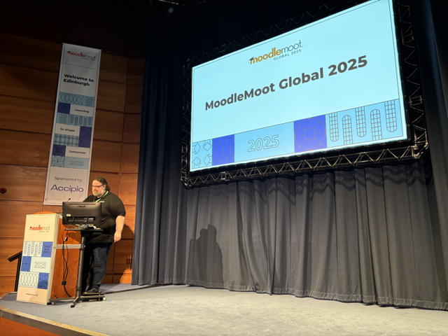 Gavin Henrick, Co-Founder of Brickfield Education Labs, is stood to the left on a small, brightly lit stage. Above, is a large screen displaying "MoodleMoot Global 2025" with the MoodleMoot logo above.