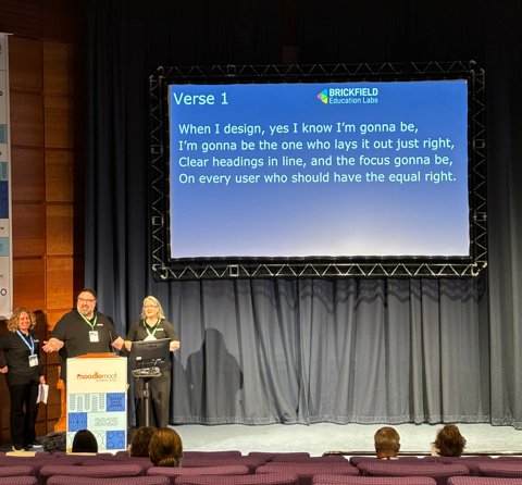 An auditorium with attendees in seated rows facing a brightly lit stage. On the stage is a large screen displaying "Verse 1" of Brickfield's version of the song 'I'm Gonna Be (500 Miles), by the Proclaimers'. The screen reads "When I design, yes I know I'm gonna be, I'm gonna be the one who lays it out just right, clear headings in line, and the focus gonna be, on every user who should have equal right". Gavin, Karen, and Laia stand on stage to the left of the screen singing along.