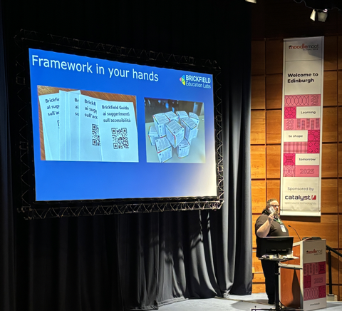An auditorium with attendees in seated rows facing a brightly lit stage. On the stage is a large screen displaying the logo "Brickfield Education Labs" with a colourful geometric design in the top right. The slide displays the title "Framework in your hands" with two images below. The image to the left represents booklets with QR codes and the image to the right represents a stack of cube shaped resources related to accessibility. Gavin, Co-Founder of Brickfield Education Labs, stands in front of the screen to the right speaking to the audience.
