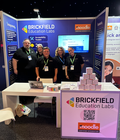 Group photo of five team members from Brickfield Education Labs, all wearing black polos with the company's logo printed in small. Behind them is a stand with the company's logo "Brickfield Education Labs" with a small geometric, colourful design. Moodle's certified integration logo is also on the stand, consisting of an orange rectangle with "Moodle" written in white writing and a graduation cap sitting on the "M" with the words certified integration below it. The booth setup consists of a TV and Brickfield promoting accessibility solutions. In front of the team is a table with a chair and a laptop. To the right of the table is another table with Brickfields logo and a QR code. Resting on the table is a 3D pyramid of Brickfields accessibility cubes.