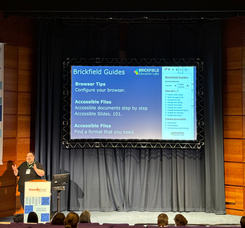 An auditorium with attendees in seated rows facing a brightly lit stage. On the stage is a large screen displaying a presentation slide titled "Brickfield Guides". The slide lists browser tips and accessible file options, such as configuring your browser, creating accessible documents and slides, and finding alternative formats. To the left of the screen, Gavin Henrick Co-Founder of Brickfield Education Labs, stands at a podium presenting.