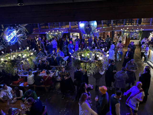 A lively costume party seen from a balcony view. Groups of people are gathered around long wooden tables and standing across the room, chatting, and holding drinks. Many attendees are dressed in themed costumes, including capes, helmets, and togas. Overhead, decorative chandeliers with hanging greenery and lights add to the atmosphere, alongside a glowing neon sign on the wall.