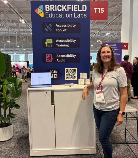 Kara Conger standing and smiling beside Brickfield Education Labs booth at Educause Annual Conference. The booth features a large vertical sign highlighting accessibility services including a toolkit, training, and audit. A laptop and promotional materials sit on the counter, and other attendees and booths are visible in the background.