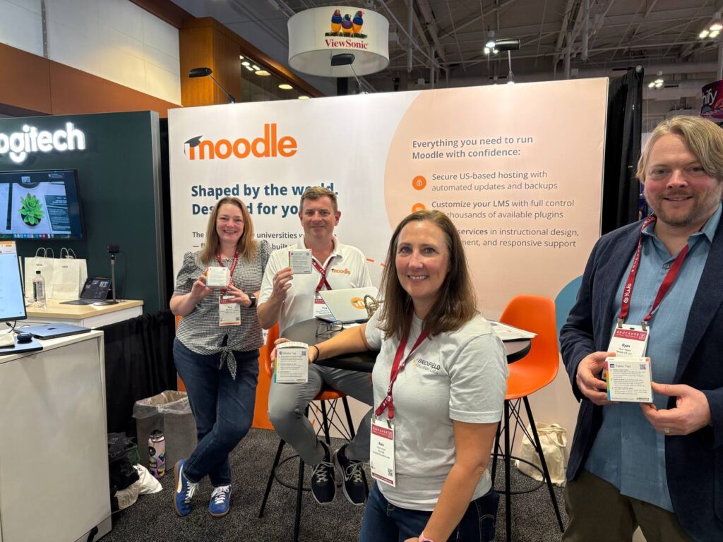 Kara Conger and the Moodle team at the Educause annual conference, smiling and holding Brickfield accessibility tip cubes. Booth signage behind them highlights Moodle logo.