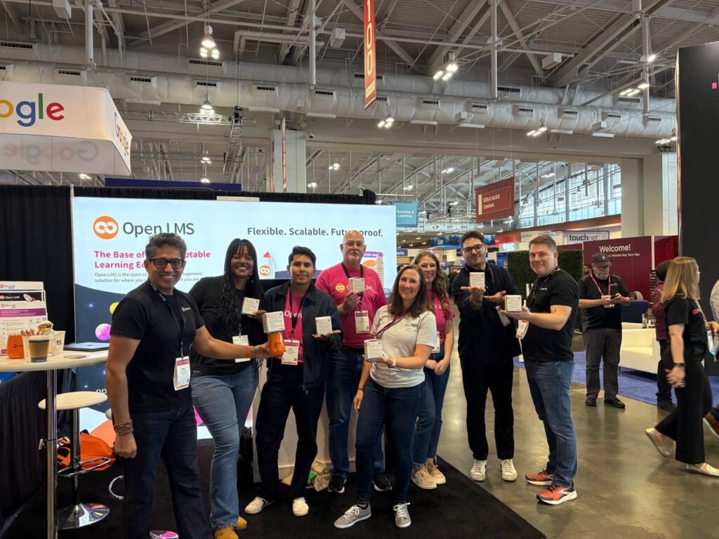 Open LMS team and Kara Conger from Brickfield Education Labs smiling in front of the Open LMS stand at Educause annual conference. Everyone is holding Brickfield's Accessibility Tip Cubes.