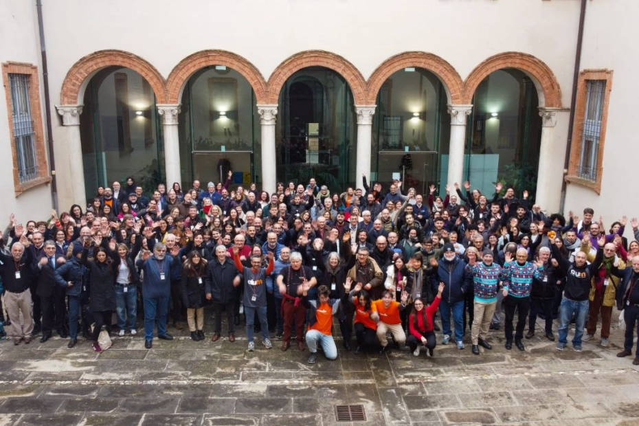 Large group of MoodleMoot Italia attendees standing in a historic courtyard in Ferrara, many waving toward the camera.