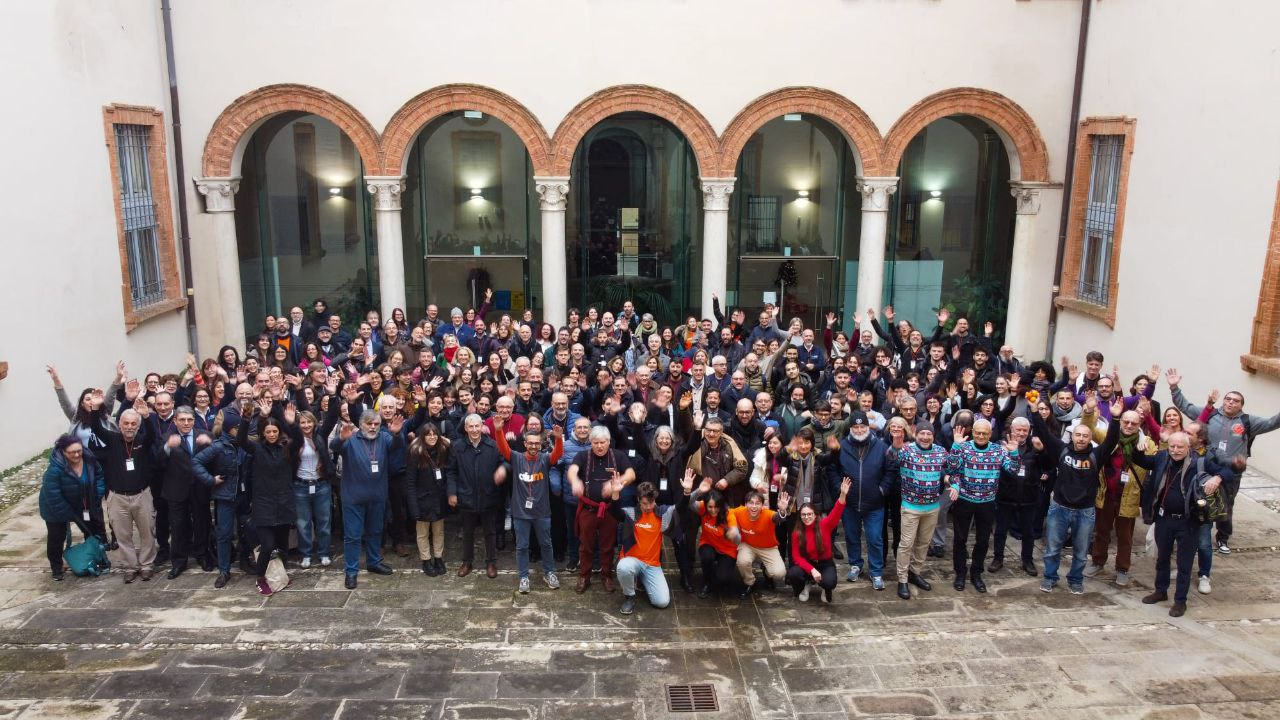 Large group of MoodleMoot Italia attendees standing in a historic courtyard in Ferrara, many waving toward the camera.