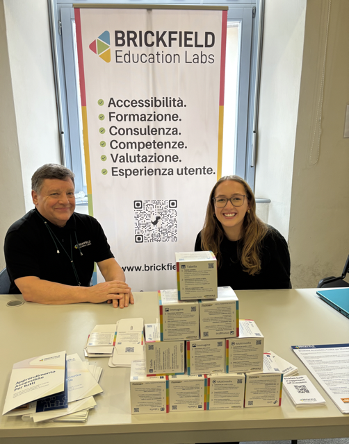 Two people sitting behind a table at Moodle Moot Italy event booth. A vertical banner behind them reads Brickfield Education Labs with Italian text about accessibility, training, and consulting. The table displays stacked product boxes and printed materials. Jerry O'Sullivan on the left wears a black, Brickfield shirt and smiles; Annabelle Snow on the right wears glasses and also wears a Brickfield t-shirt.