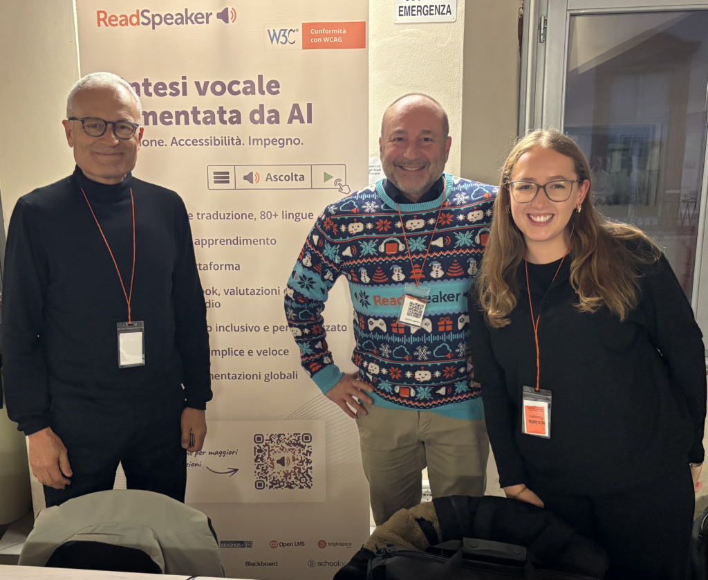 Two team members from ReadSpeaker and one from Brickfield Education Labs standing in front of a ReadSpeaker banner. The man on the left wear's glasses and a dark jumper, the man in the centre wears a patterned ReadSpeaker jumper, and the woman on the right smiles wearing glasses and a dark top. They all wear conference badges. The banner behind them shows the ReadSpeaker logo and text about AI-powered speech and accessibility features.