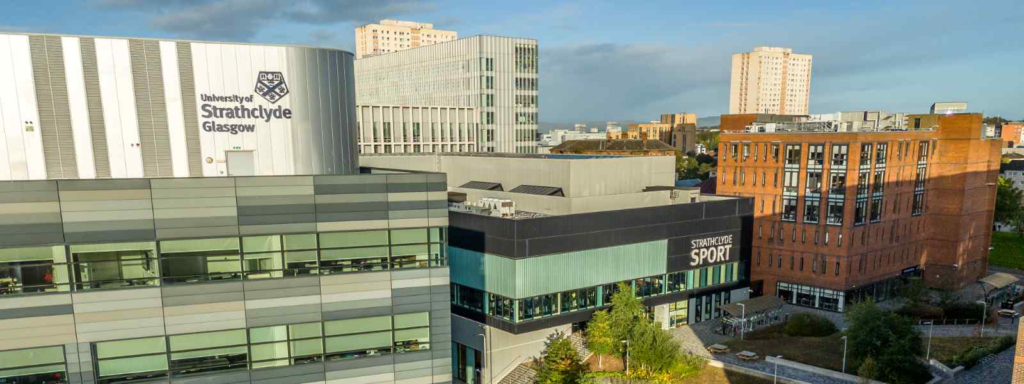 A view of the University of Strathclyde in Glasgow, showing several modern buildings including the Strathclyde sport centre with large windows. To the left a tall silver building with the university’s name and crest, and to the right of the sport centre is a large red-brick building. High rise city buildings are visible in the background.