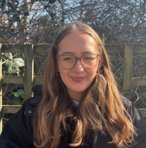 A woman with long light-brown hair and wears glasses, standing outdoors.