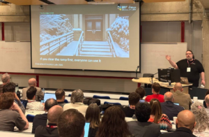 Gavin Henrick presents at MoodleMoot Canada, gesturing toward a projection screen showing a slide titled "The Paradigm Shift" from Brickfield Education Labs. The slide features a snow-covered building entrance with a ramp and stairs, captioned "If you clear the ramp first, everyone can use it.