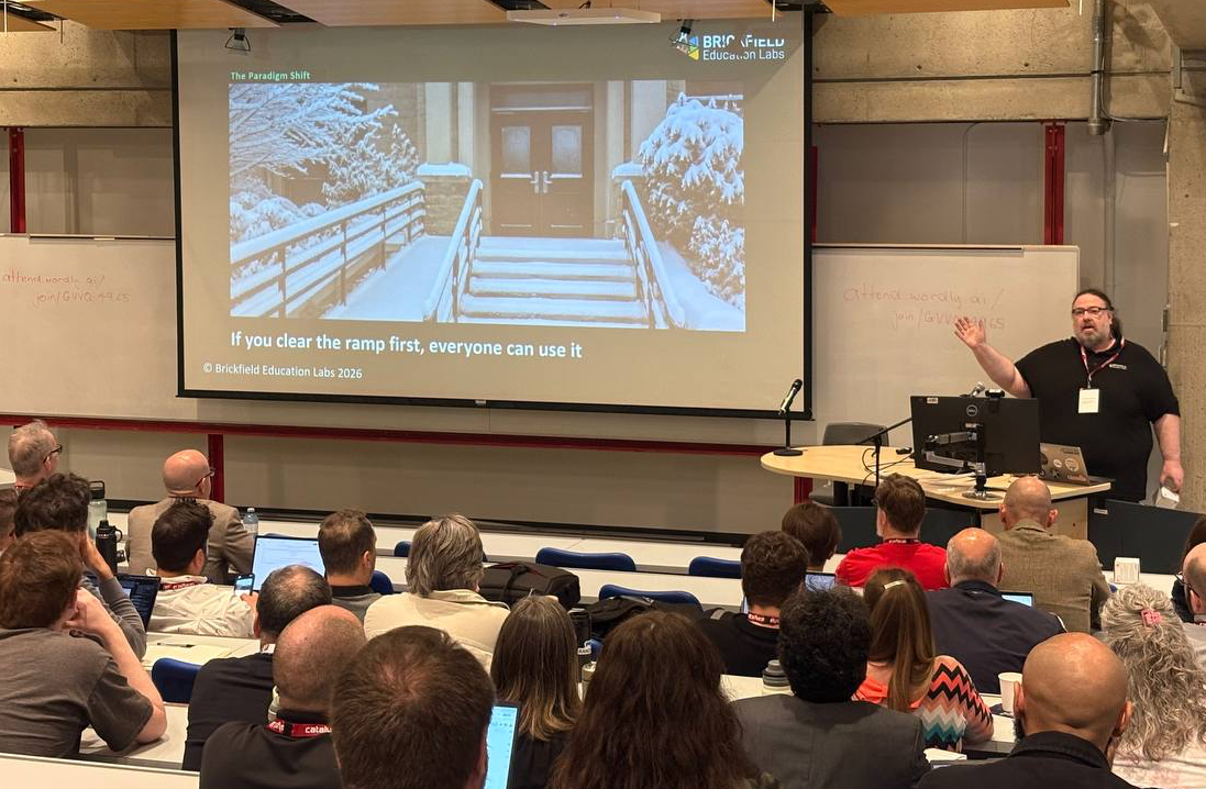Gavin Henrick presents at MoodleMoot Canada, gesturing toward a projection screen showing a slide titled "The Paradigm Shift" from Brickfield Education Labs. The slide features a snow-covered building entrance with a ramp and stairs, captioned "If you clear the ramp first, everyone can use it.