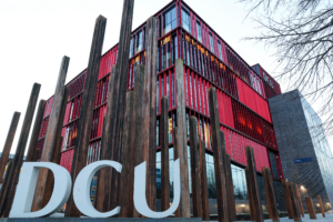 The Polaris Building at Dublin City University’s Glasnevin Campus, with its red vertical fins, behind large white DCU letters and tall timber posts.