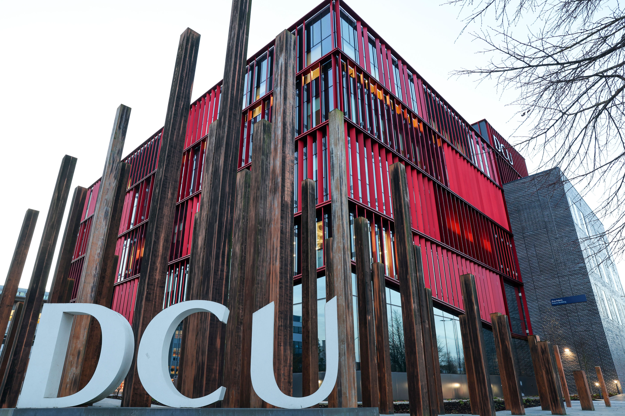 The Polaris Building at Dublin City University’s Glasnevin Campus, with its red vertical fins, behind large white DCU letters and tall timber posts.