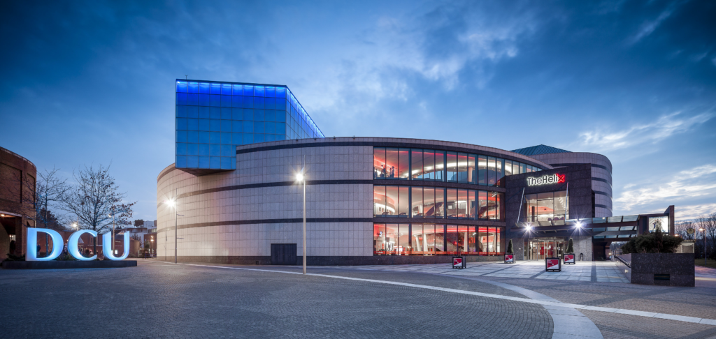 The Helix building on DCU's Glasnevin Campus at dusk, lit up blue and red, with an illuminated DCU sign in front.